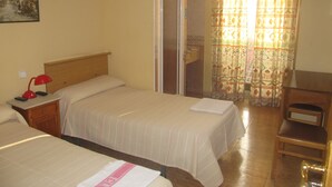 A hotel room with two beds, a nightstand with a lamp, a wooden chair, and a shower area. - Pensión Mollo (Madrid)