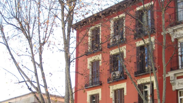 A red building with multiple windows and balconies, a visible entrance, and a street corner with pedestrians. - Pensión Mollo (Madrid)