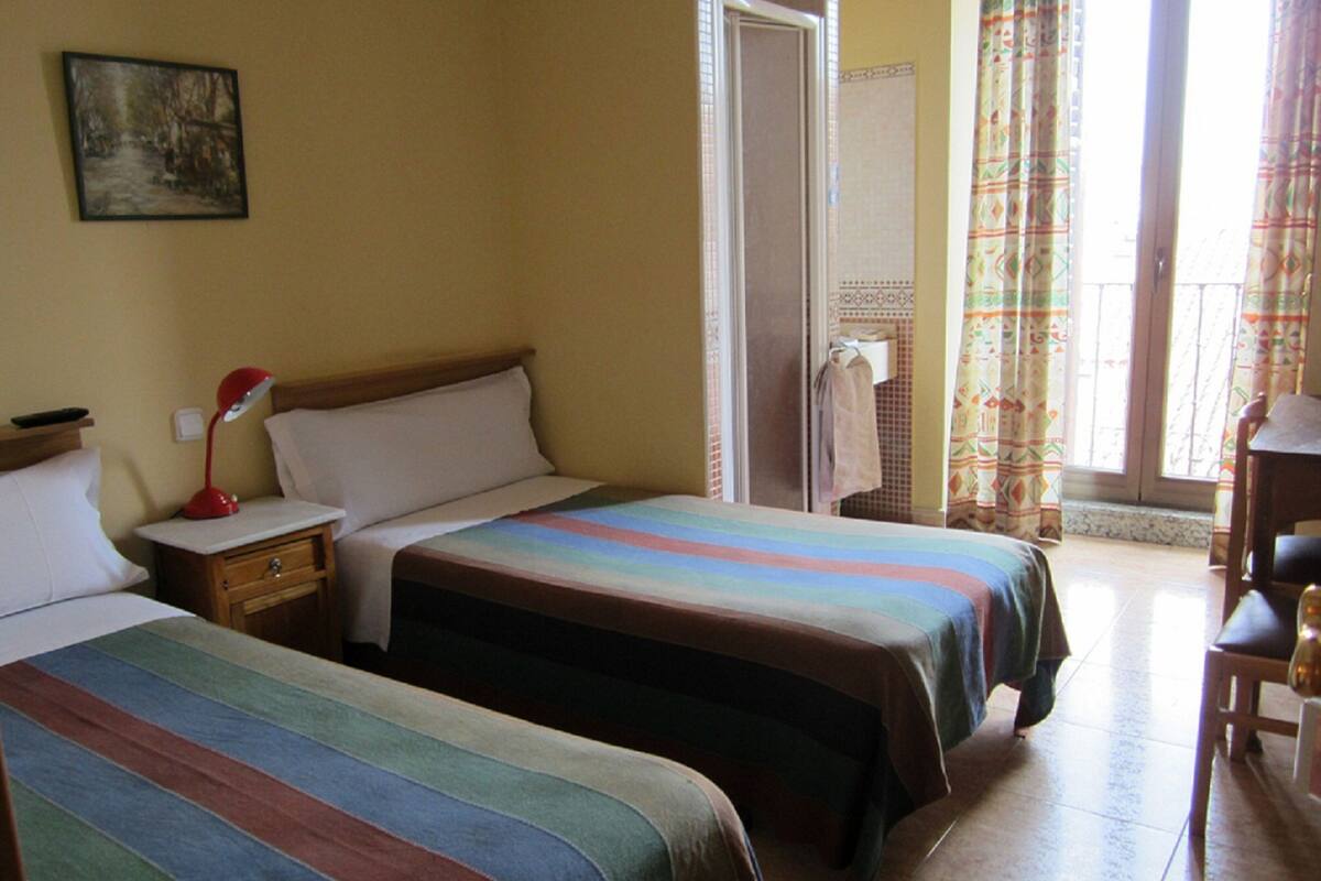 Habitación de hotel con dos camas, una mesita de noche, una lámpara roja, un cuadro en la pared y una ventana con cortinas.