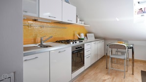 A modern kitchen with white cabinets, a gas stove, and a small dining area with a chair. - Italianway   - Alessi (Milan)