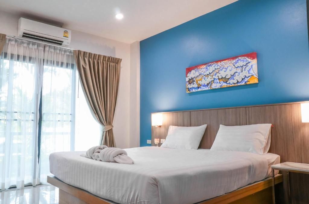 A hotel room with a bed, curtains, an air conditioning unit, a painting on the wall, and a nightstand with a lamp.