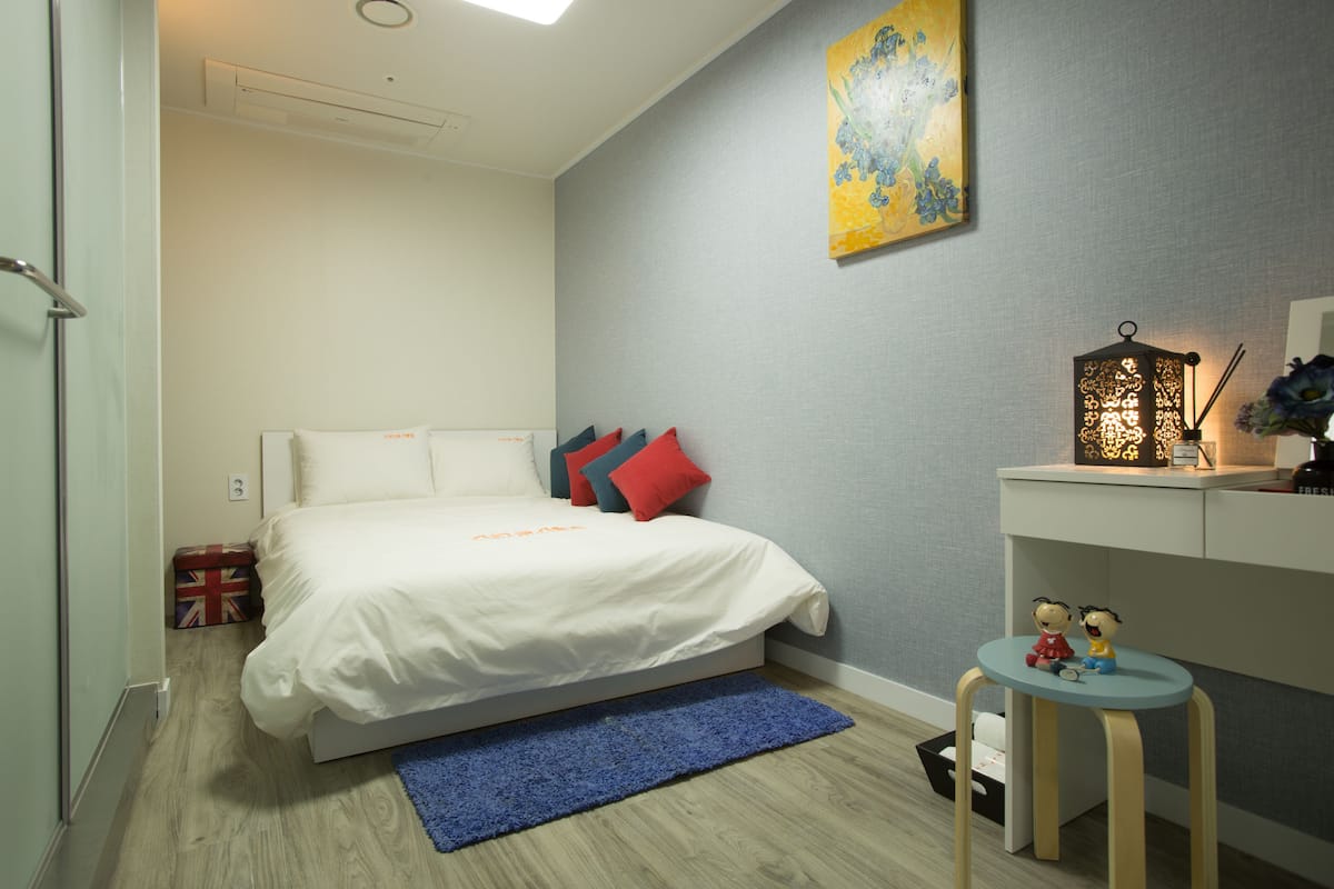 Een slaapkamer met een bed, kussens, een blauw vloerkleed, een bureau met een lamp en een schilderij aan de muur.