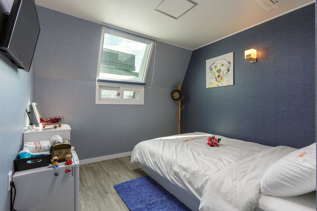 Een slaapkamer met een bed, een raam, een wandlamp en een klein bureau met een lamp en een rode speelgoedauto.