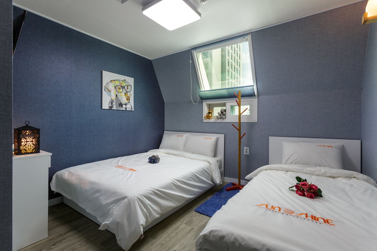 Een hotelkamer met twee bedden, een raam met uitzicht op gebouwen, een nachtkastje met een lamp en een kunstwerk aan de muur.