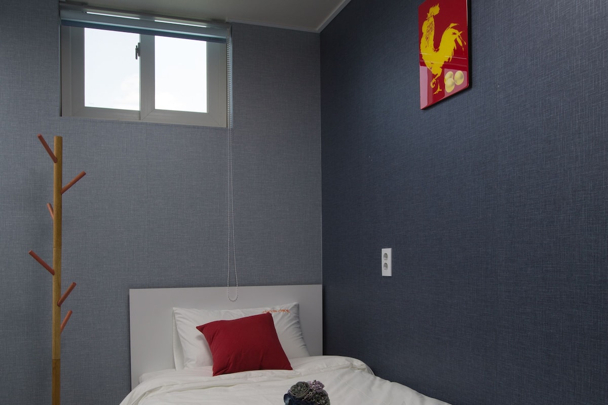 Een slaapkamer met een bed, een rood kussen, een wit kussen, een blauw vloerkleed, een houten kapstok, een raam en een schilderij aan de muur.
