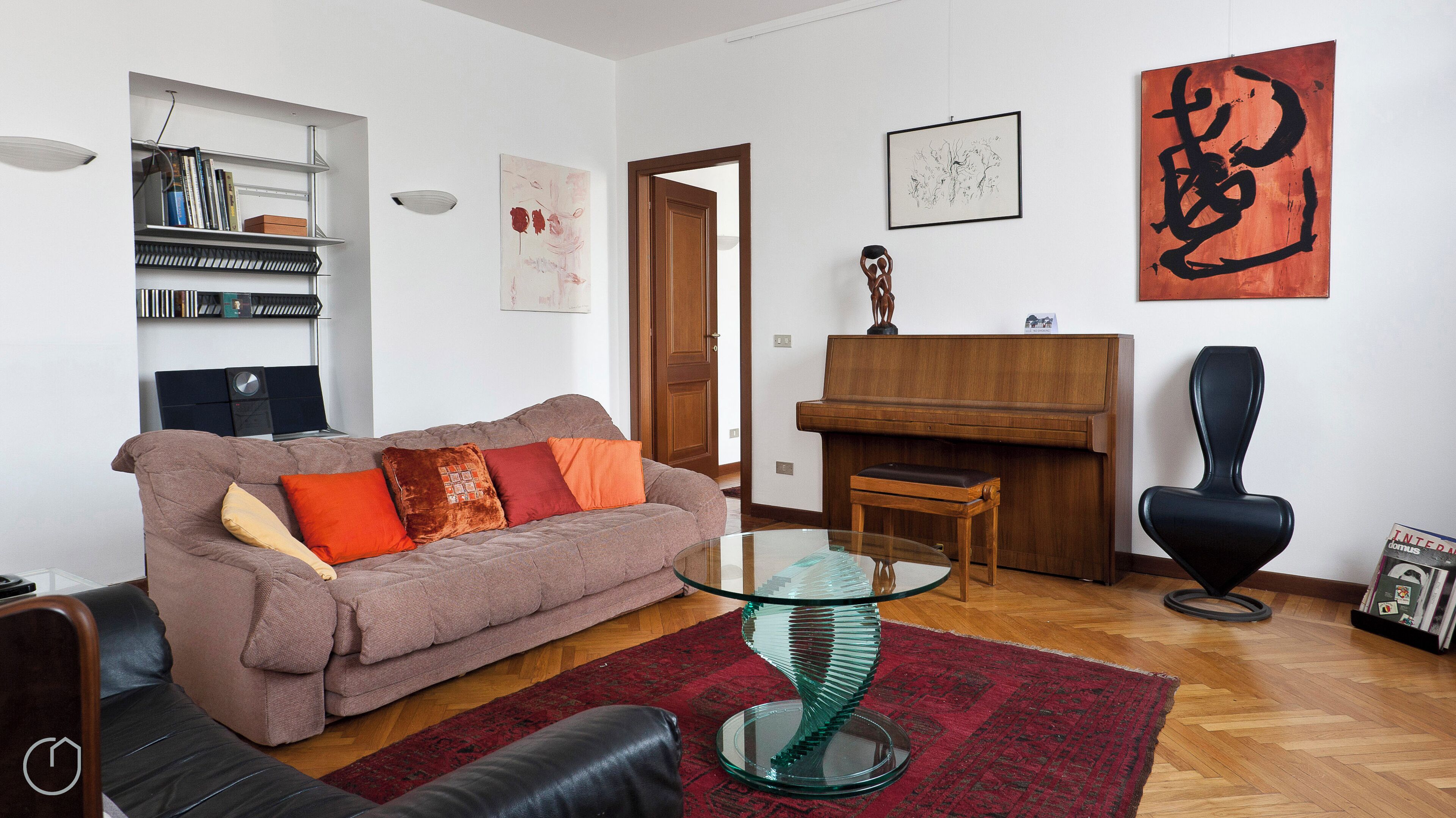 Una sala de estar con un sofá, una mesa de centro de cristal, un tapete rojo, un piano, una silla negra y una estantería.