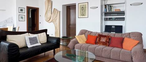 A living room with a black leather sofa, a brown sofa with colorful cushions, a glass coffee table, and a bookshelf with books and decorative items.