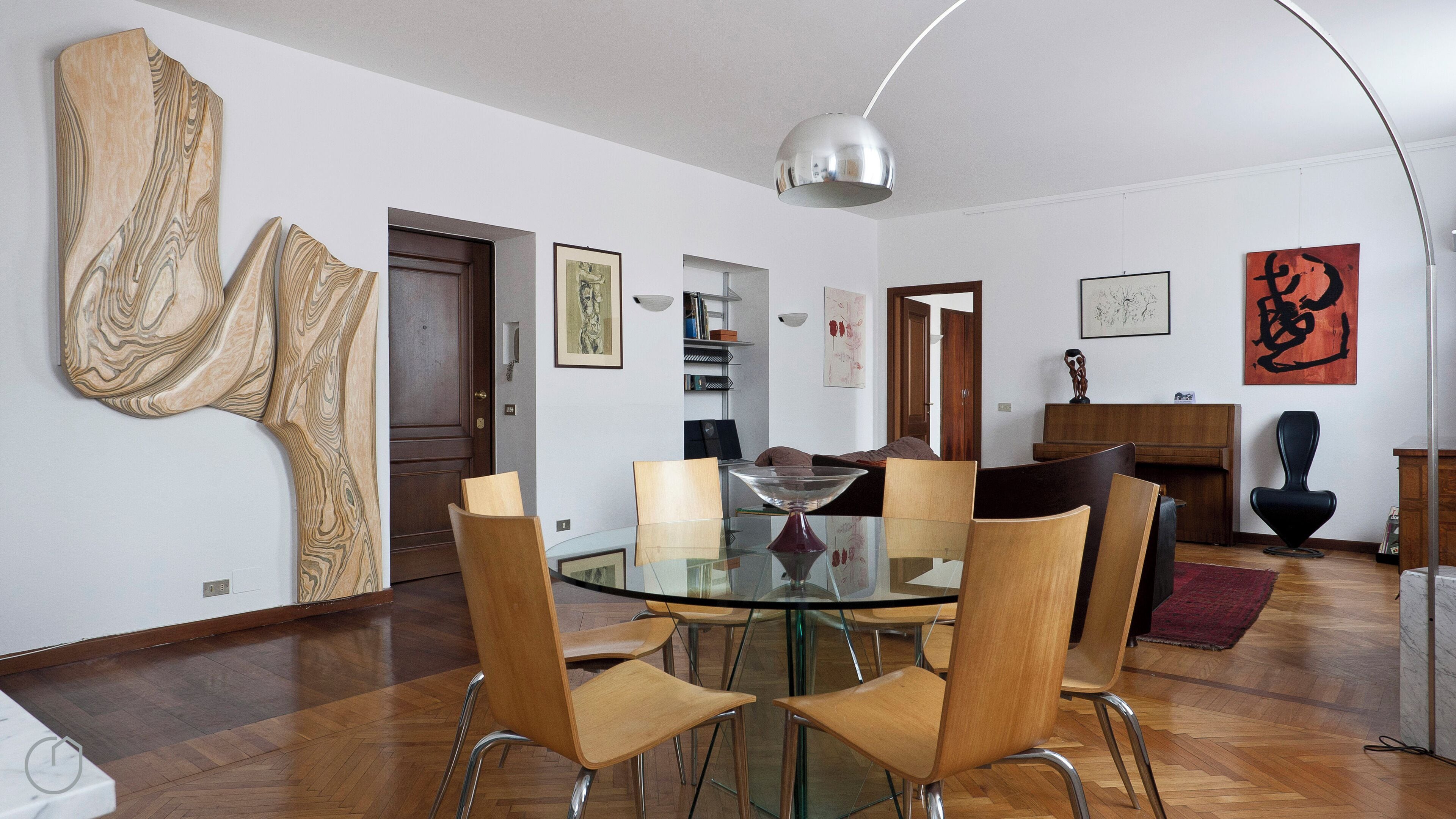 A modern dining area with a glass table and wooden chairs, a piano, and abstract art on the walls.