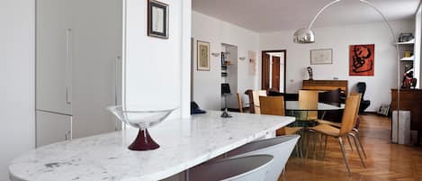 A modern kitchen with a marble countertop, a dining area with wooden chairs, and a visible hallway leading to a living space.