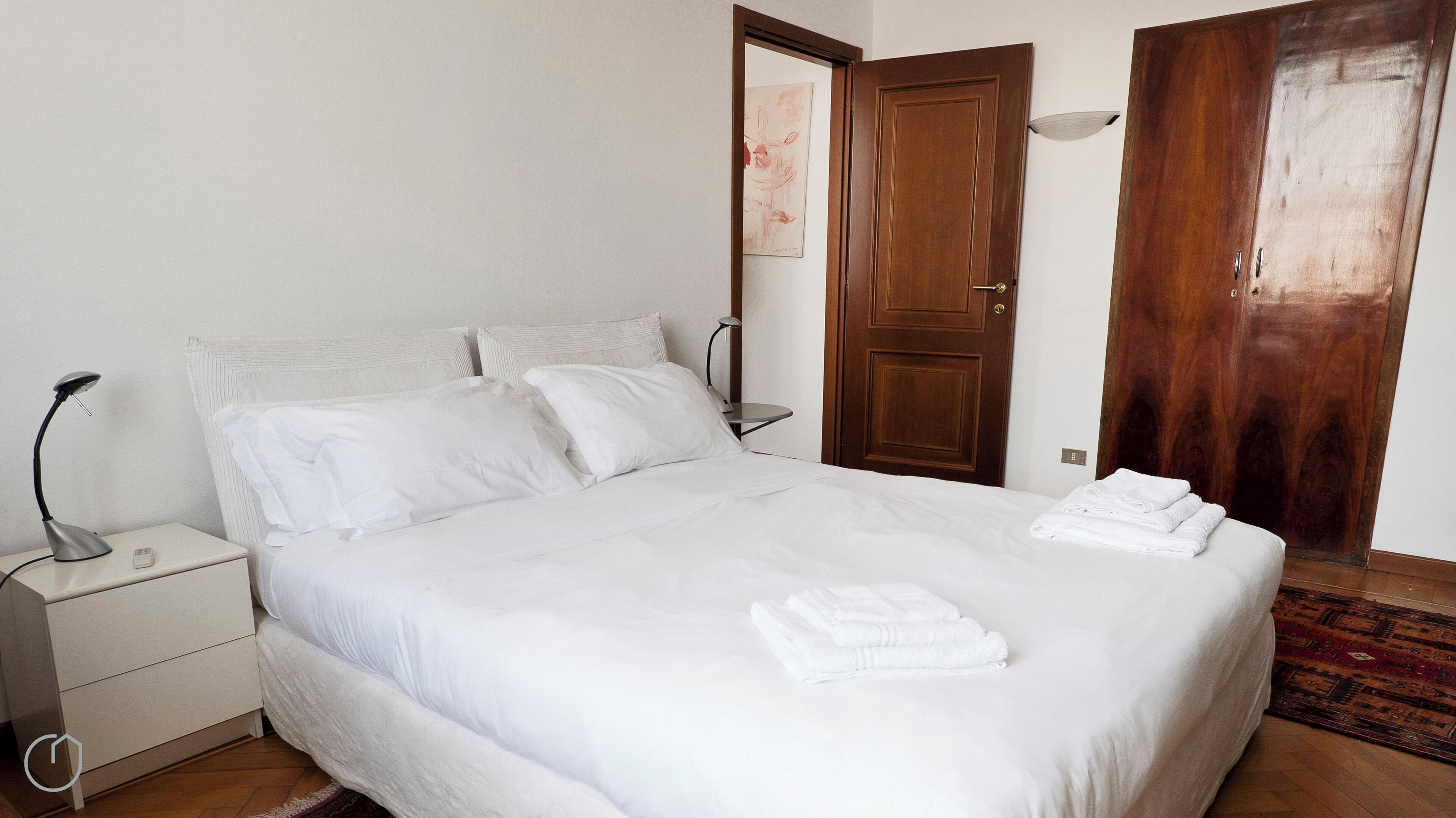 Una cama bien hecha con sábanas blancas, una mesita de noche con lámpara y una puerta de madera al fondo.