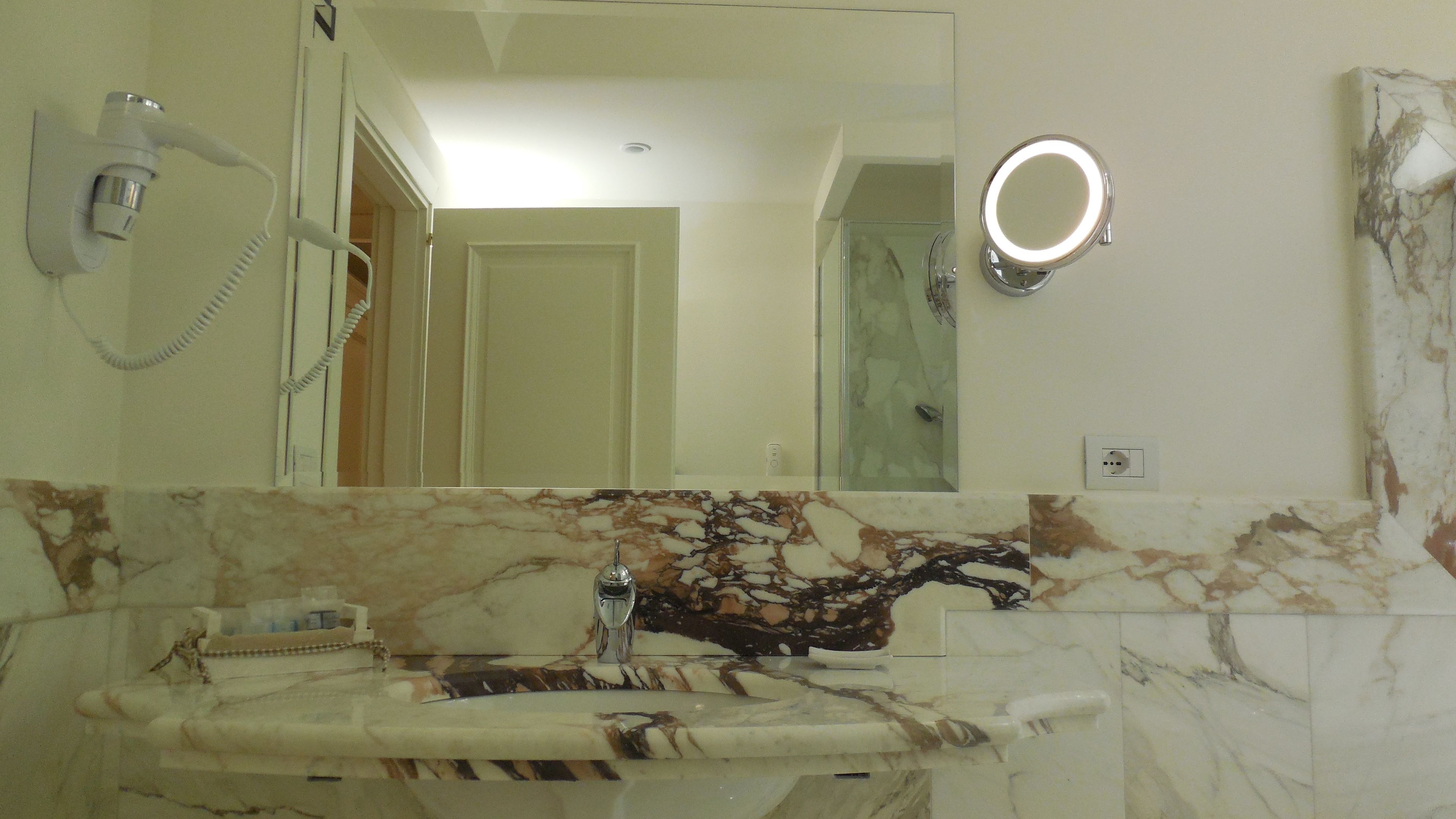 A bathroom with a marble sink, a handheld hair dryer, a mirror with a light, and a shower area.