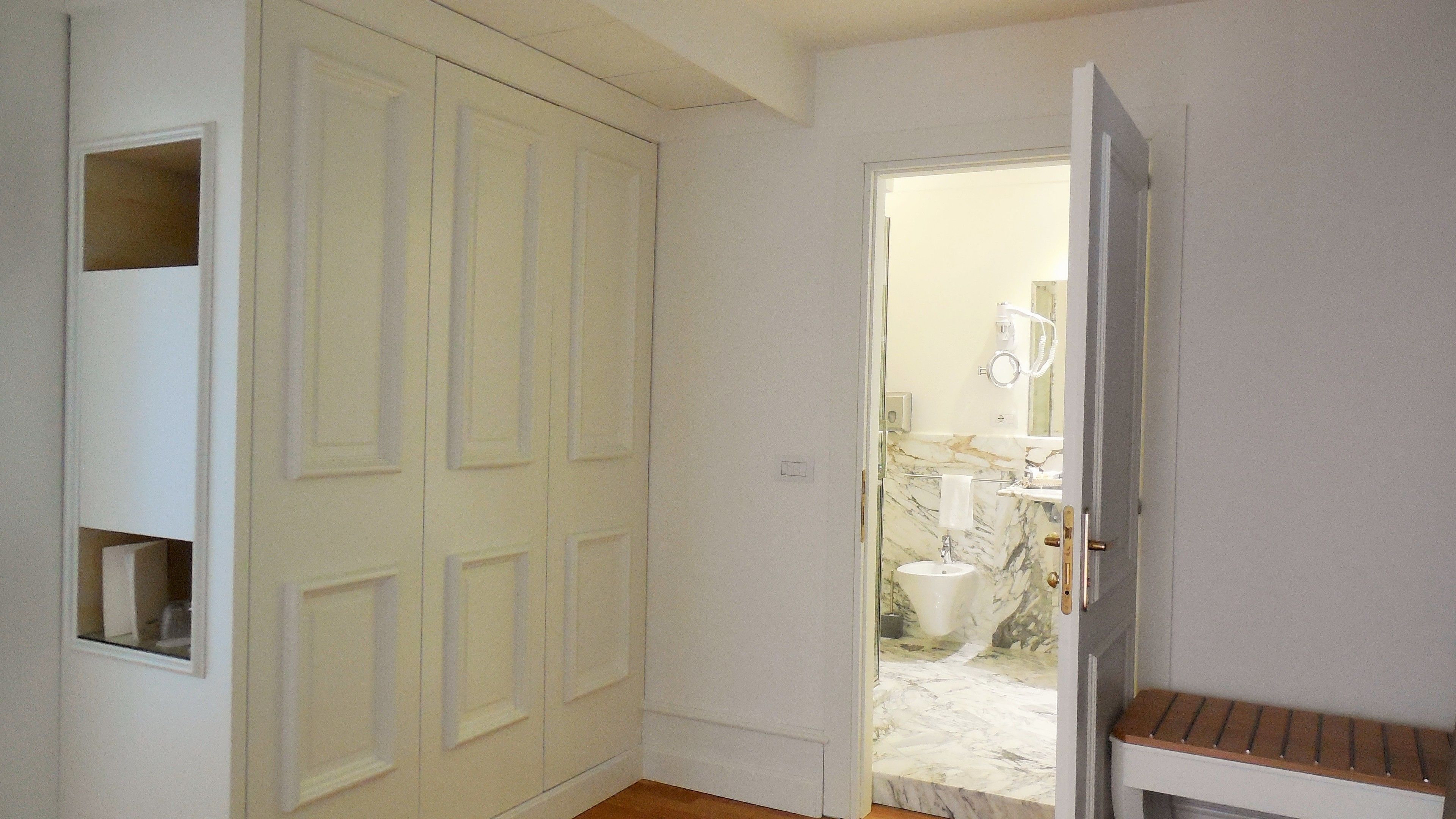 Un pasillo con una puerta cerrada, un banco y un baño visible a través de una puerta abierta.