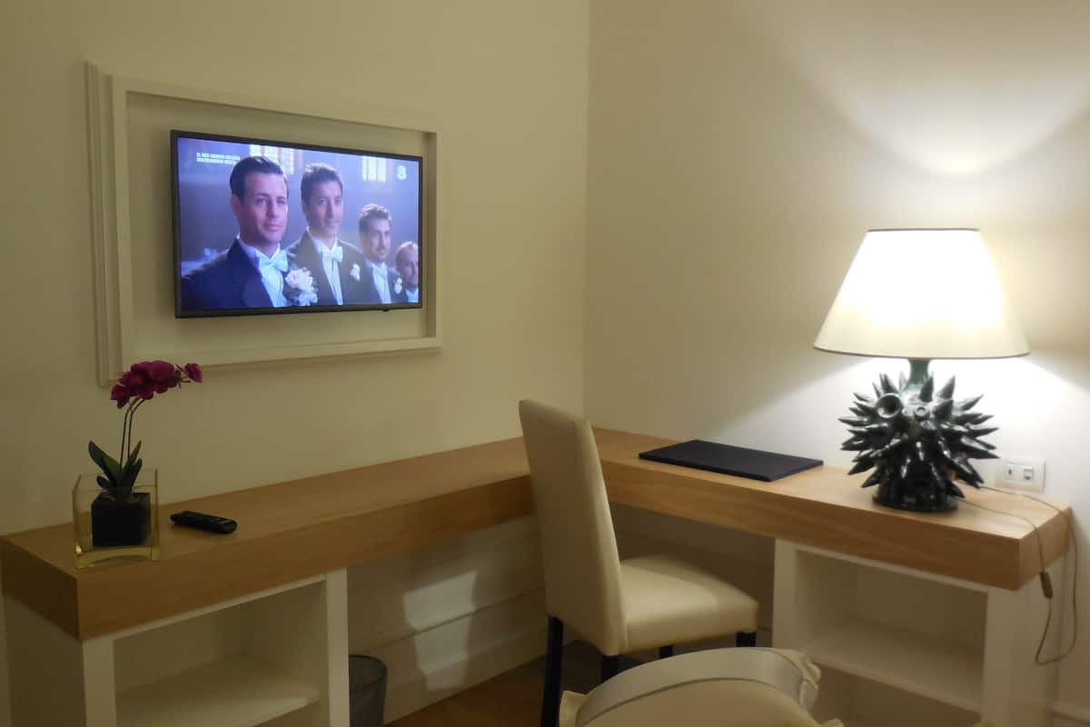 A hotel room with a desk, a TV mounted on the wall, a lamp, and a potted plant.