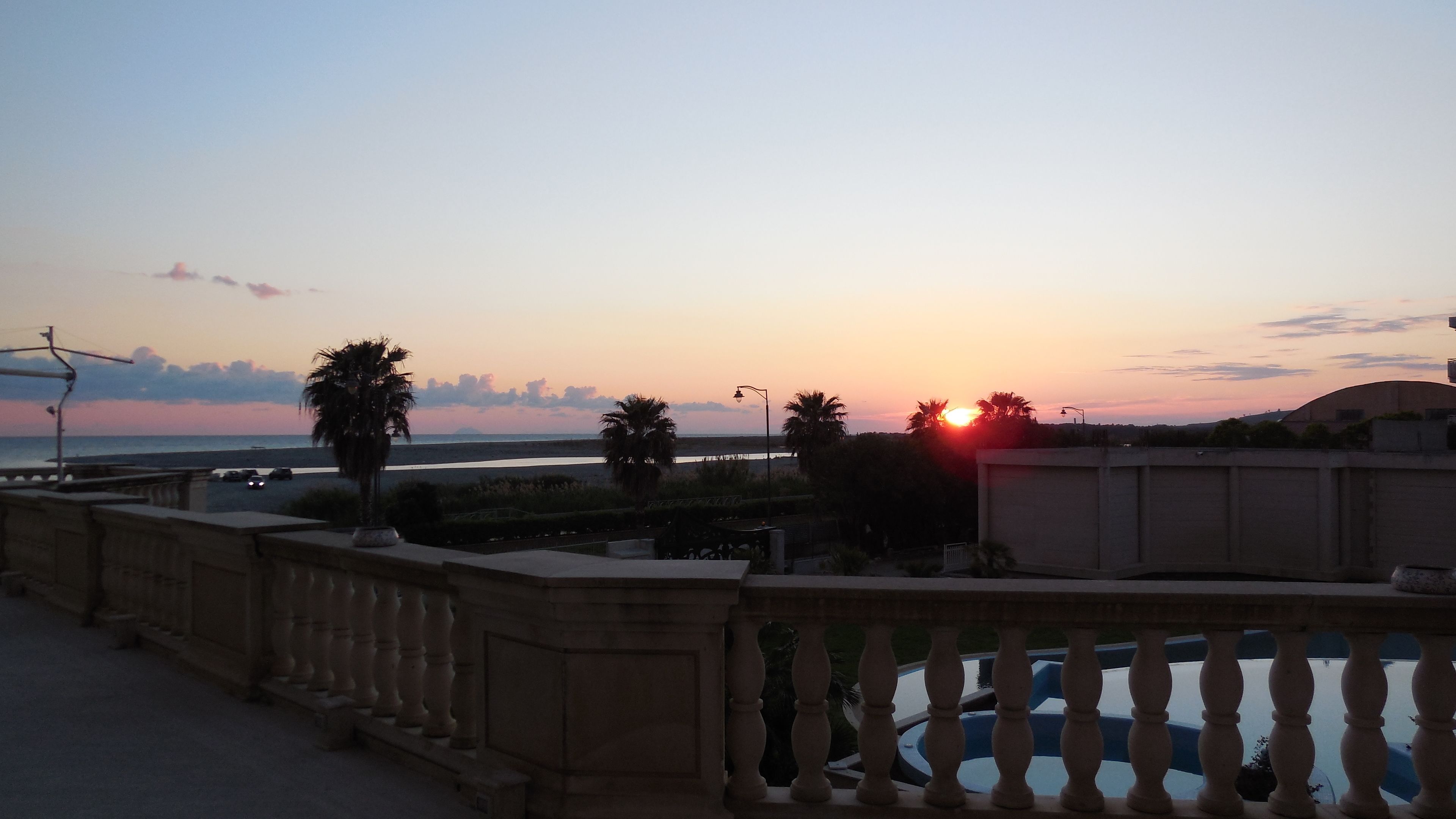 Zonsondergang vanaf een balkon met uitzicht op een strand, palmbomen en een weg.