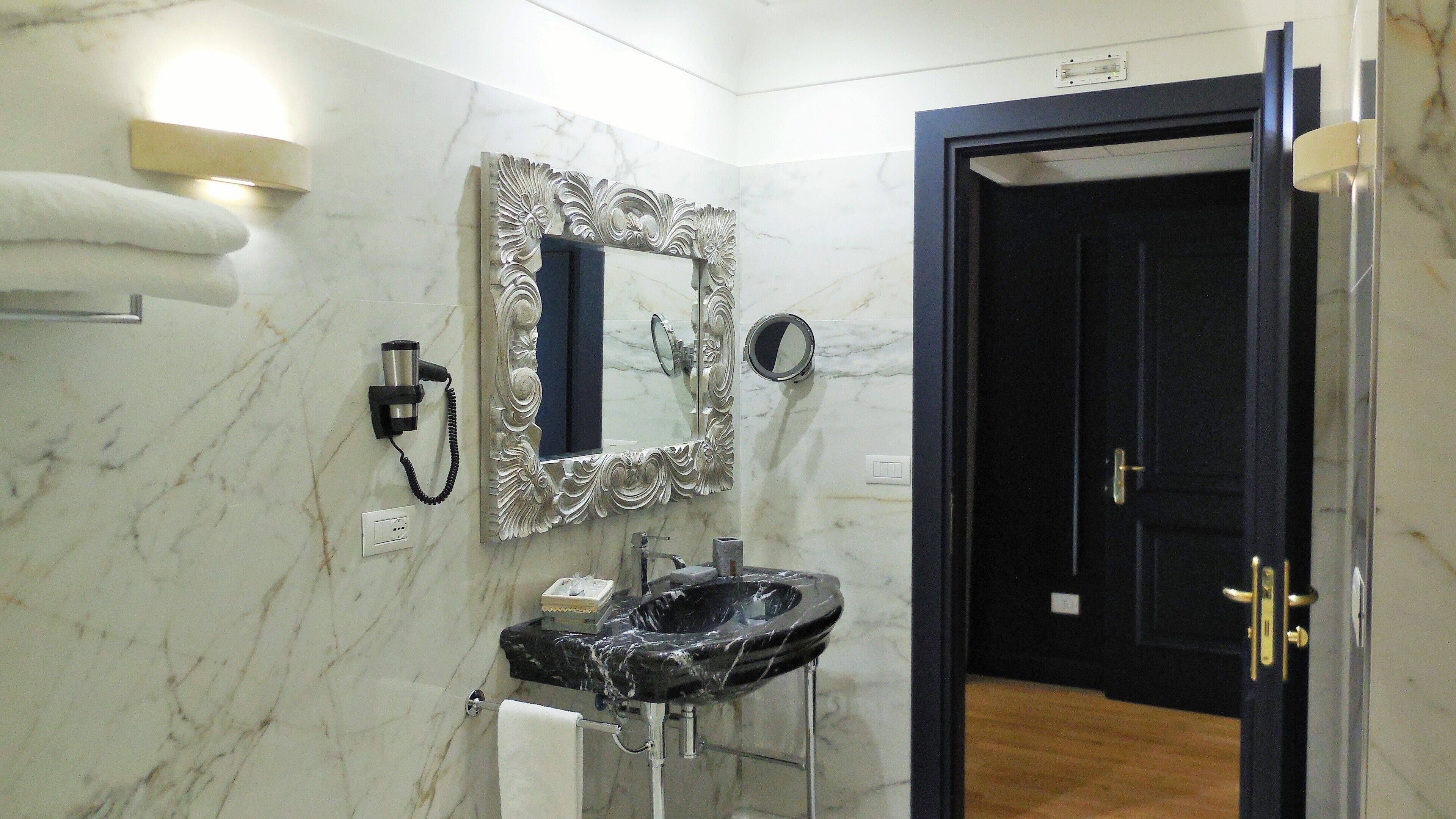 A bathroom with a marble wall, a black sink, a mirror with an ornate frame, a hairdryer, and a door.