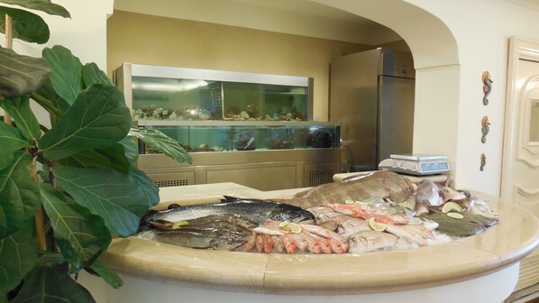A seafood display with various fish and seafood on ice, a fish tank in the background, and a kitchen area with a refrigerator and scale. - Hotel Marechiaro (Gizzeria)