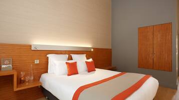 A hotel room with a wooden headboard, a bed with white and orange bedding, a bedside table with a glass, and a wooden wardrobe.