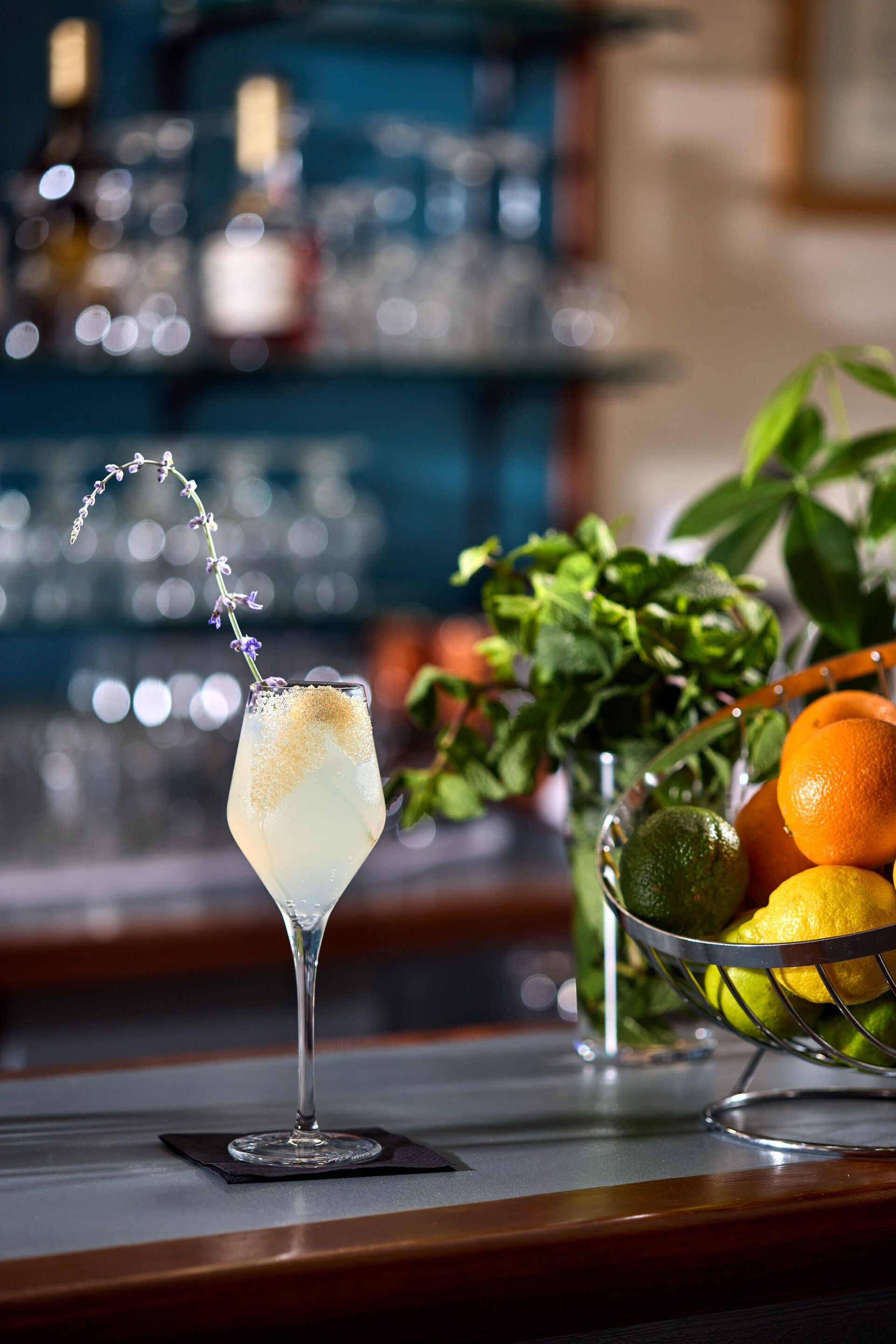 Un verre à cocktail orné d’une branche sur le bord, un panier de fruits contenant des oranges et des citrons verts, et un bar en arrière-plan.