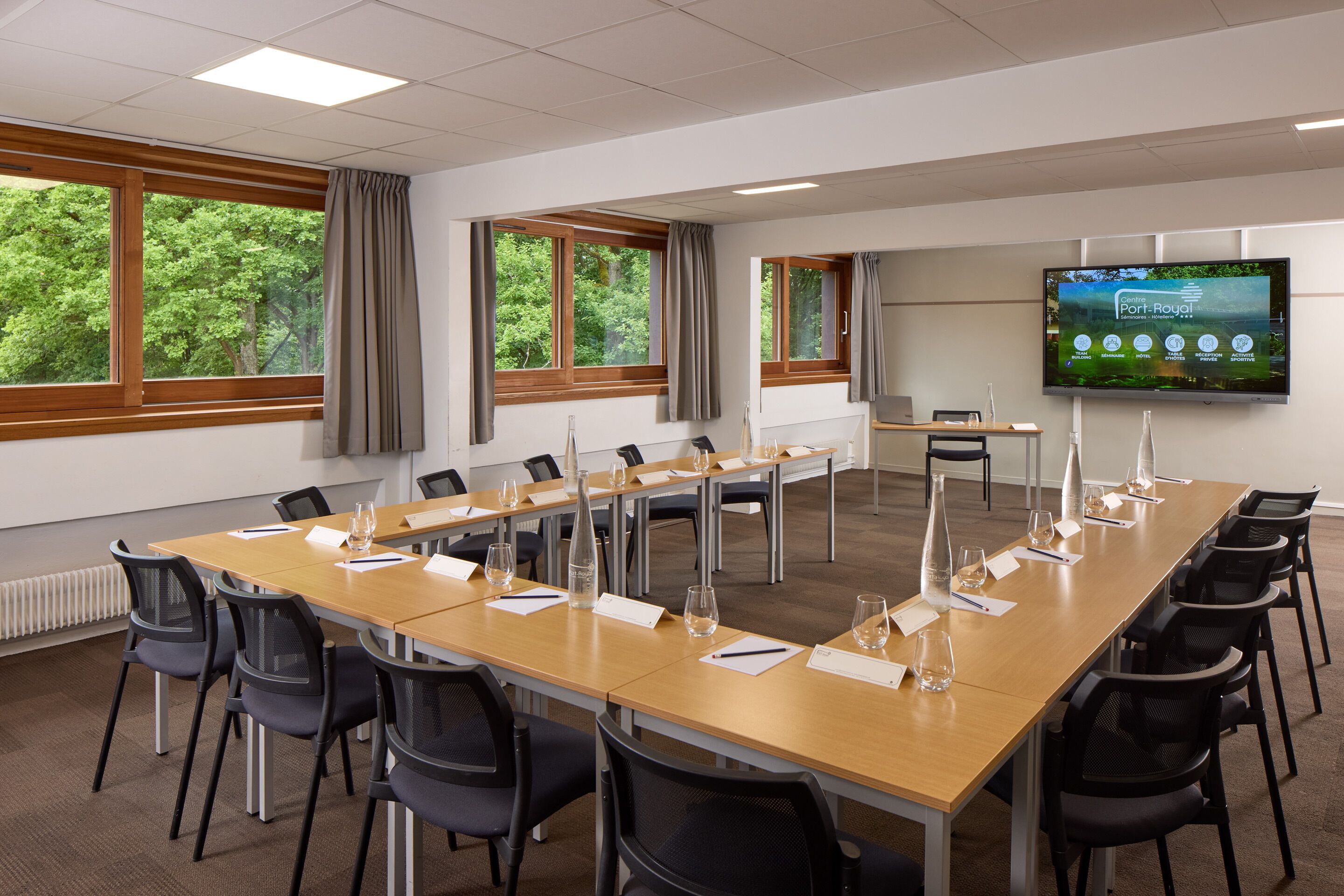 Un salón de conferencias con una larga mesa, sillas, una pantalla de proyector y ventanas con cortinas.