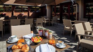 An outdoor dining area with wicker chairs, a table set with croissants, fruit, and beverages, and a view of the building in the background. - Hôtel Centre Port-Royal (Saint-Lambert)