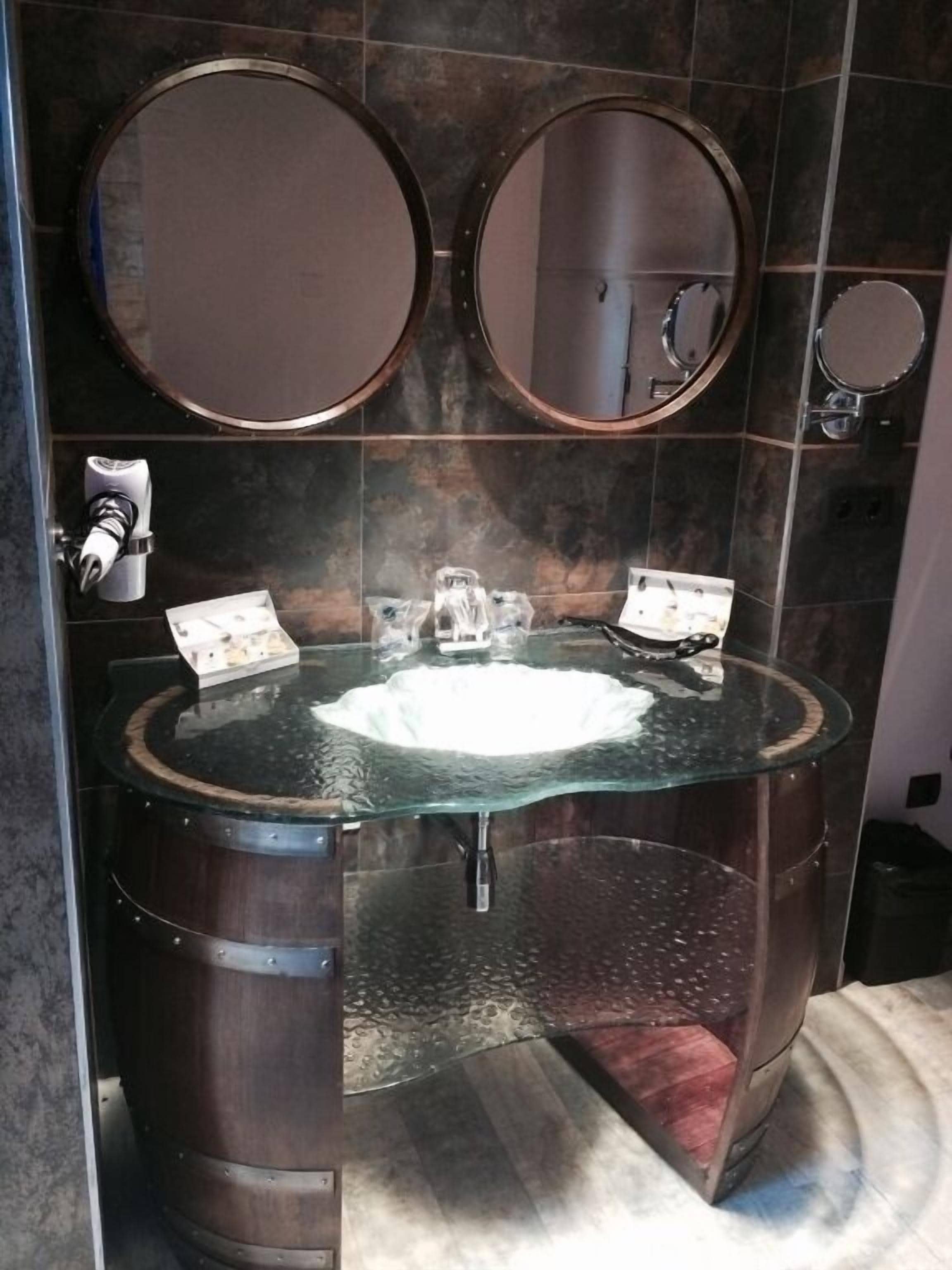 Un baño con un lavabo de vidrio redondo, dos espejos y un dispensador de toallas.