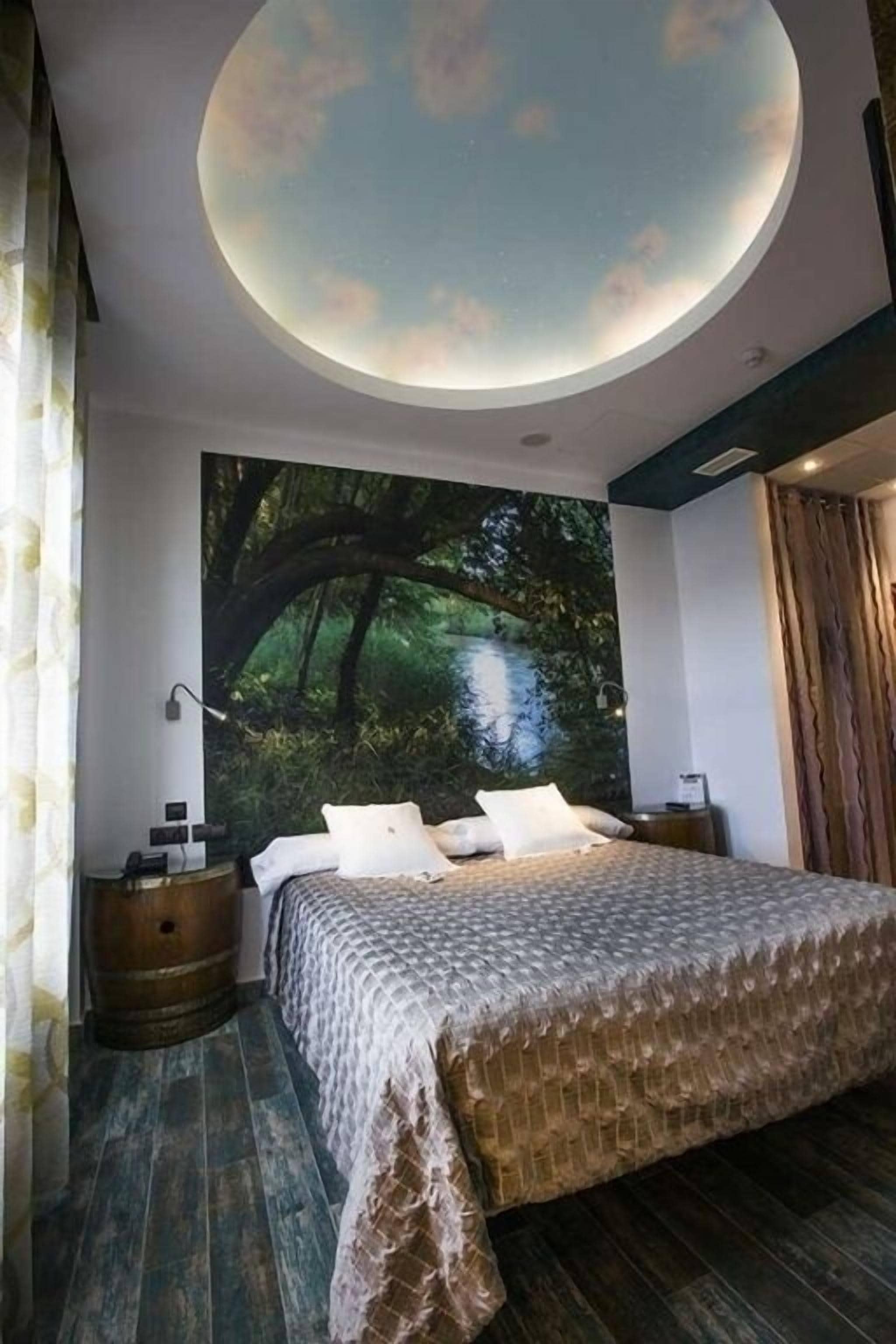 Un dormitori amb un òcul de bou circular, un llit amb un estovat estampat, una tauleta de nit i un mural gran amb motius de bosc.