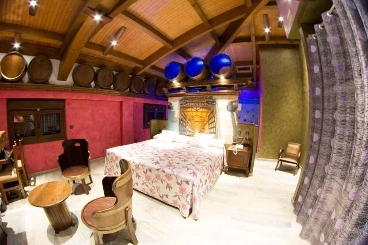 A room with a bed, wooden chairs, a table, a minibar, and a wall with wooden barrels.