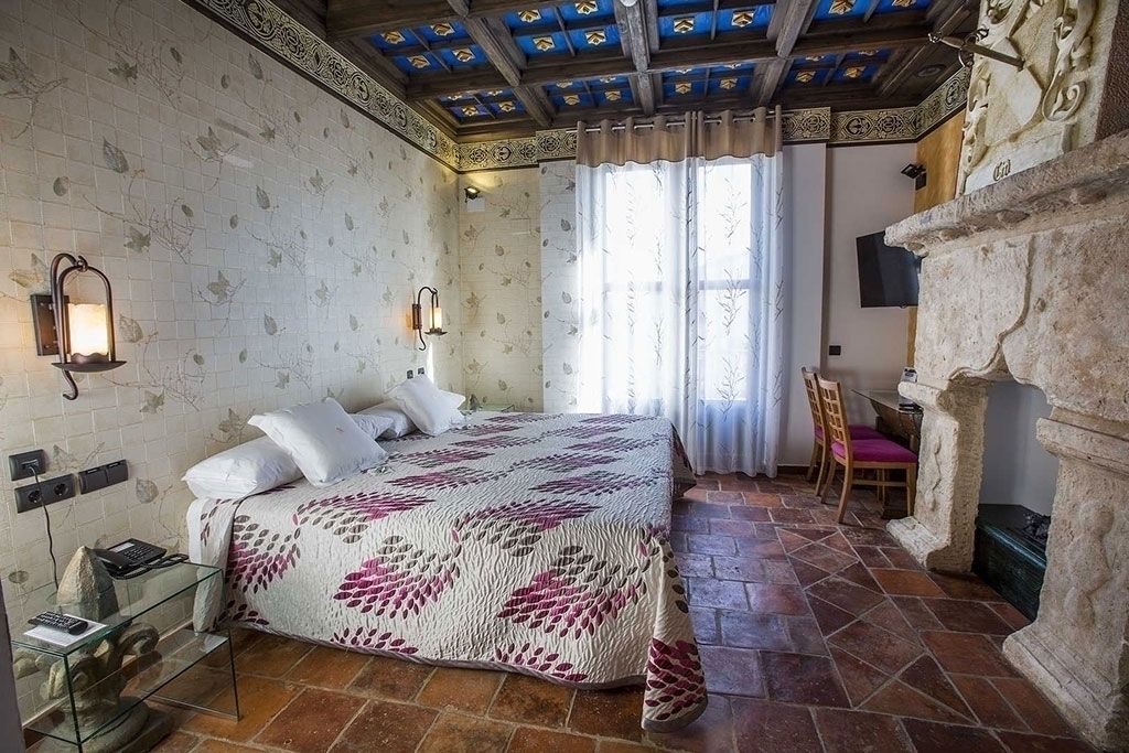 Un dormitorio con cama, una silla, chimenea de piedra y suelo de baldosas.