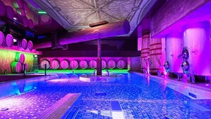 Una amplia piscina interior con suelo de baldosas, rodeada de barriles de madera y un techo con diseño geométrico.