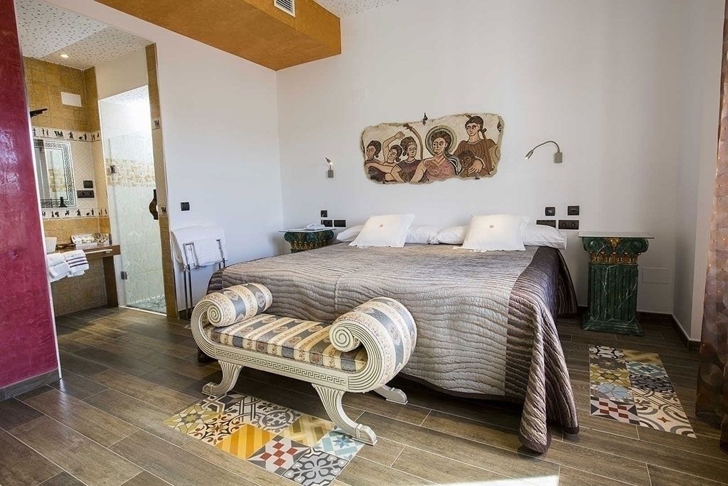 Un dormitorio con una cama grande, una banca, un cuadro en la pared y un baño visible a través de una puerta abierta.