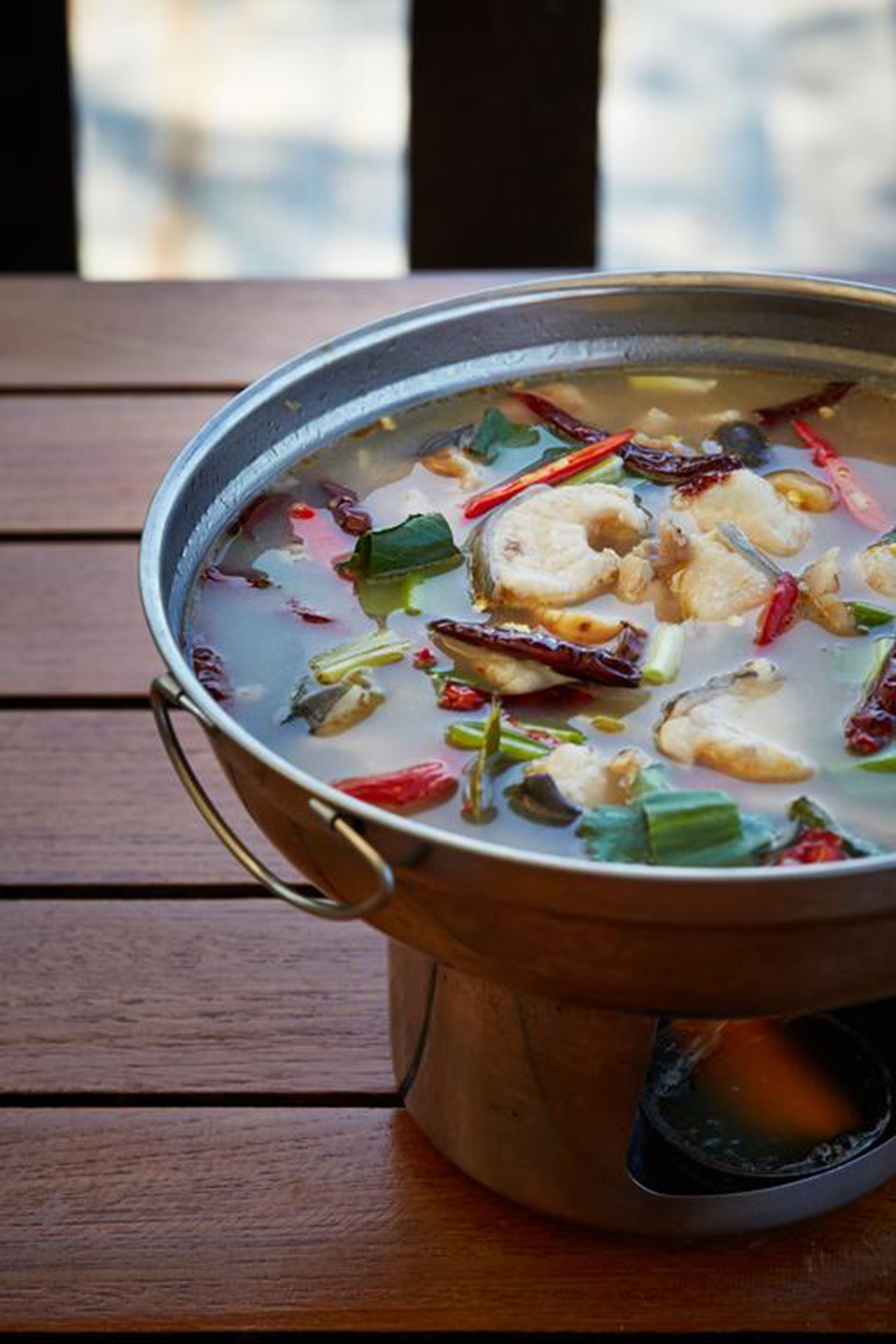 A pot of soup with visible ingredients like fish, green onions, and red chili peppers on a wooden table.