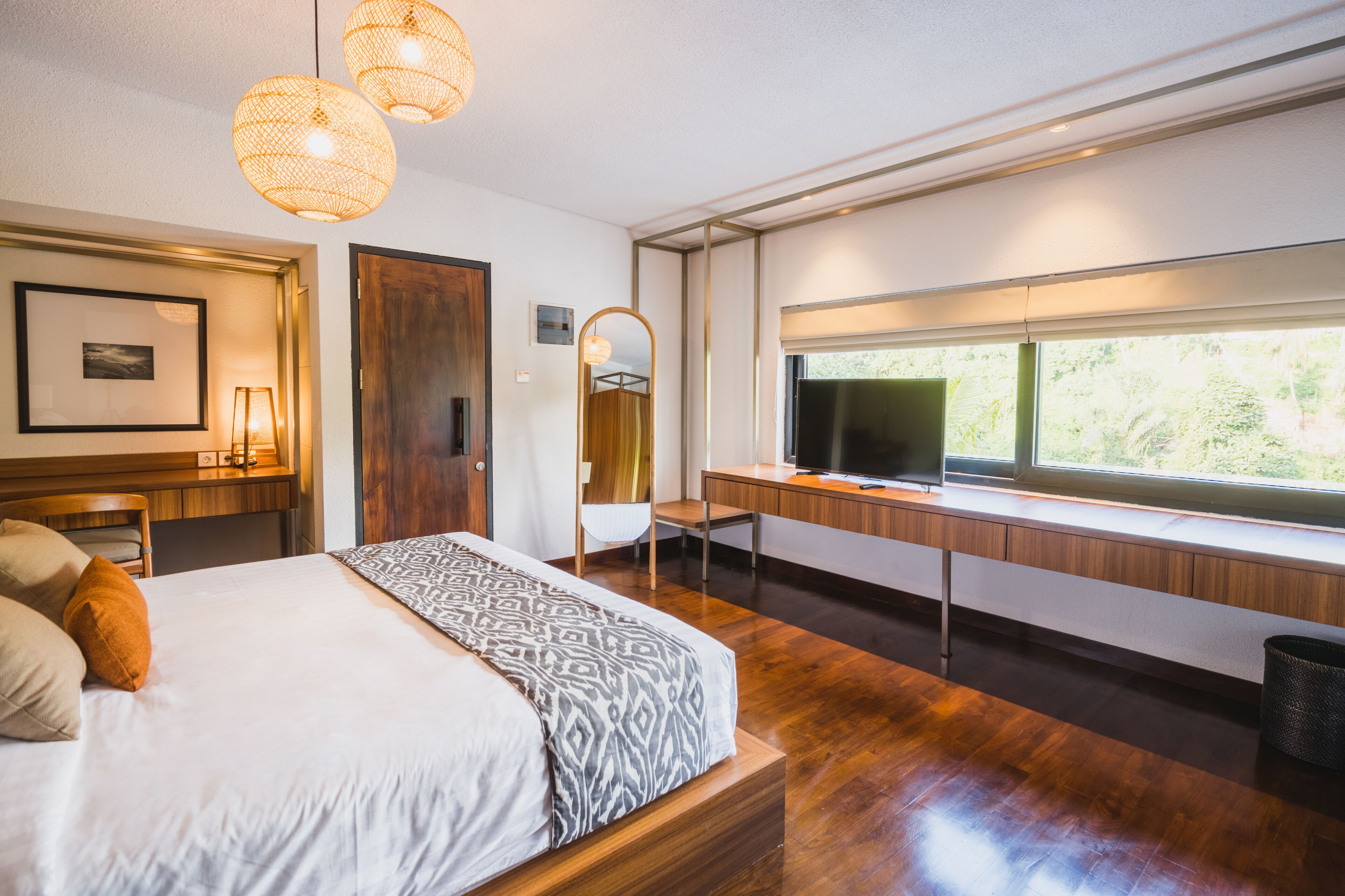 A modern hotel room with a large bed, a desk with a computer, a TV, and a window with a view of greenery.