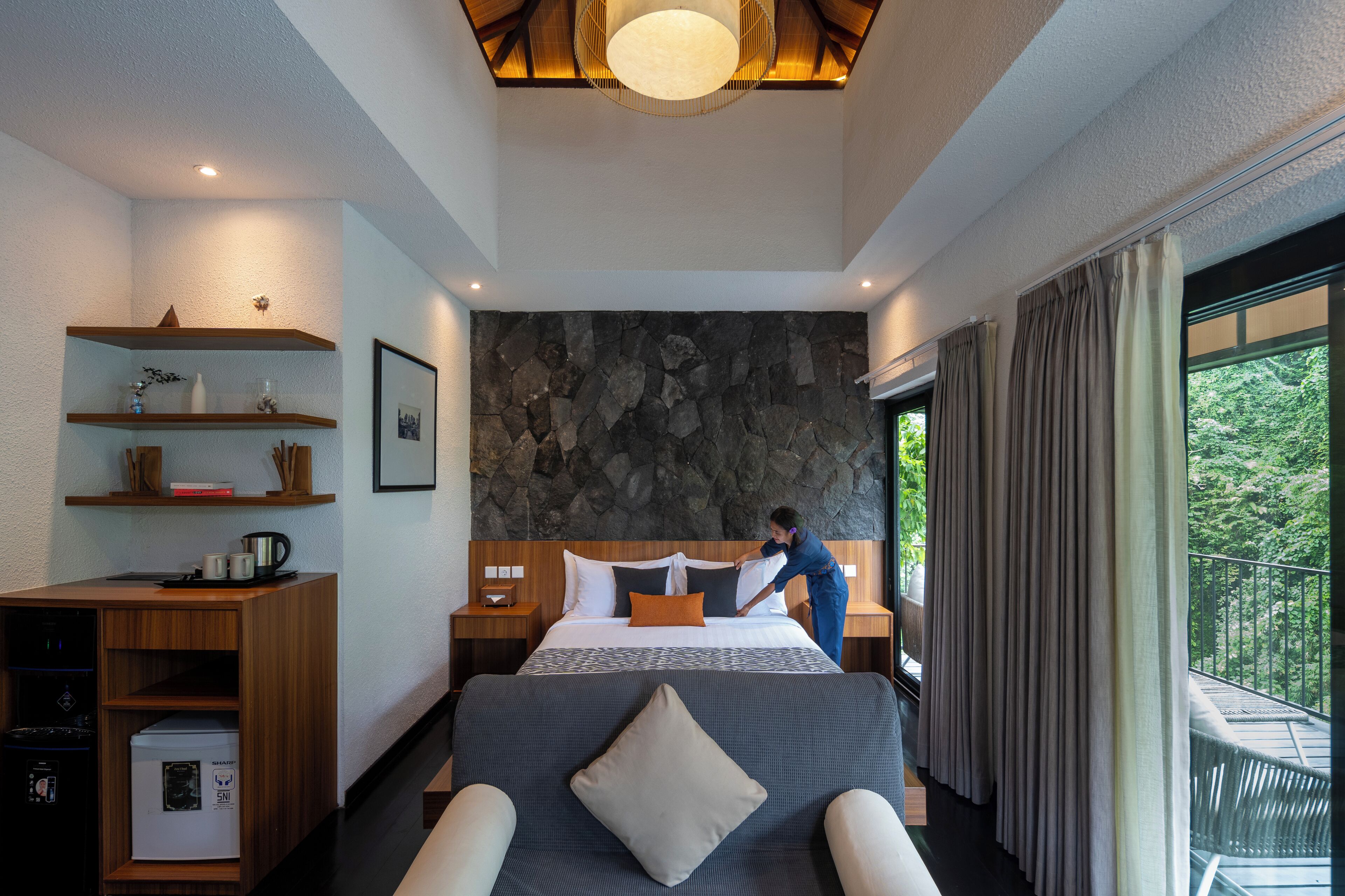 A modern hotel room with a large bed, a stone accent wall, a wooden desk with a coffee maker, and a balcony with curtains.