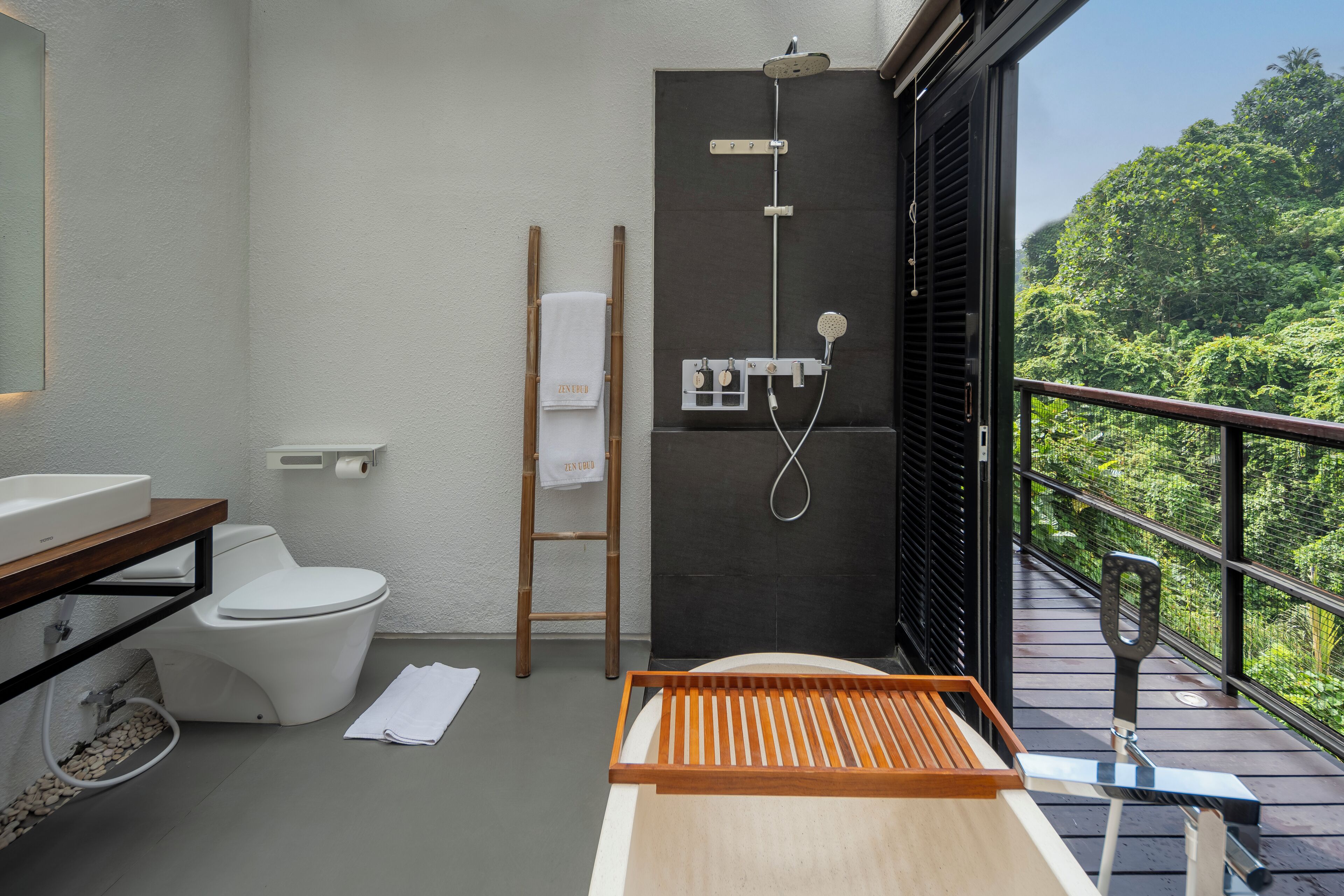 A modern bathroom with a toilet, sink, and shower. There is a wooden ladder and a towel rack. The room has a balcony with a view of greenery.