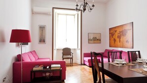 Una sala de estar con un sofá rosa, una mesa de comedor dispuesta con vasos y platos, una lámpara de pie roja y una araña.