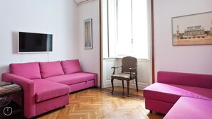 Una sala de estar moderna con dos sofás rosados, un suelo de madera en espiga, un televisor montado en la pared y un cuadro con la imagen de un edificio.