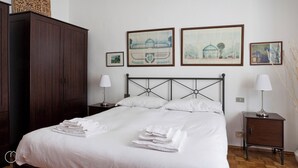 Una cama bien hecha con sábanas blancas y toallas dobladas, un armario de madera oscura y una mesita de noche con una lámpara. Hay cuadros enmarcados en la pared sobre la cama.