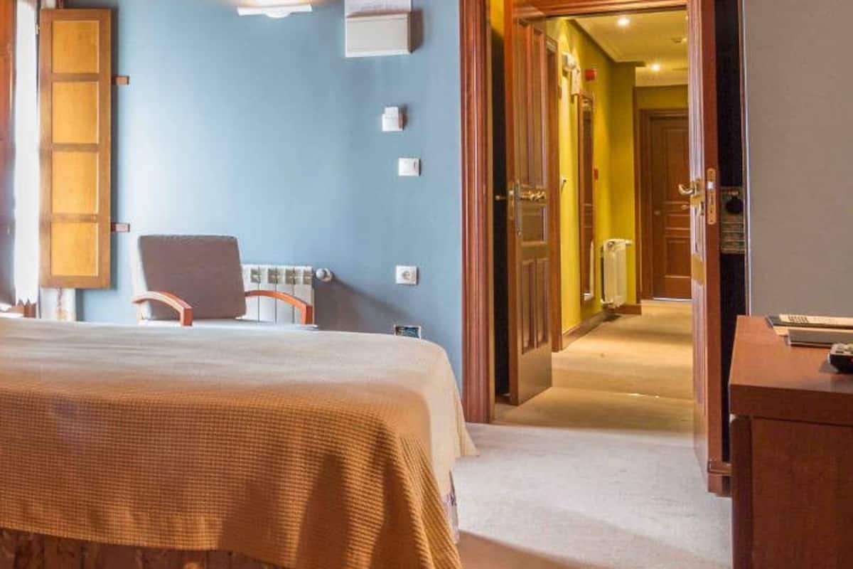 A hotel room with a bed, bedside table, and a nightstand. There is a mirror on the wall and a door leading to another room.