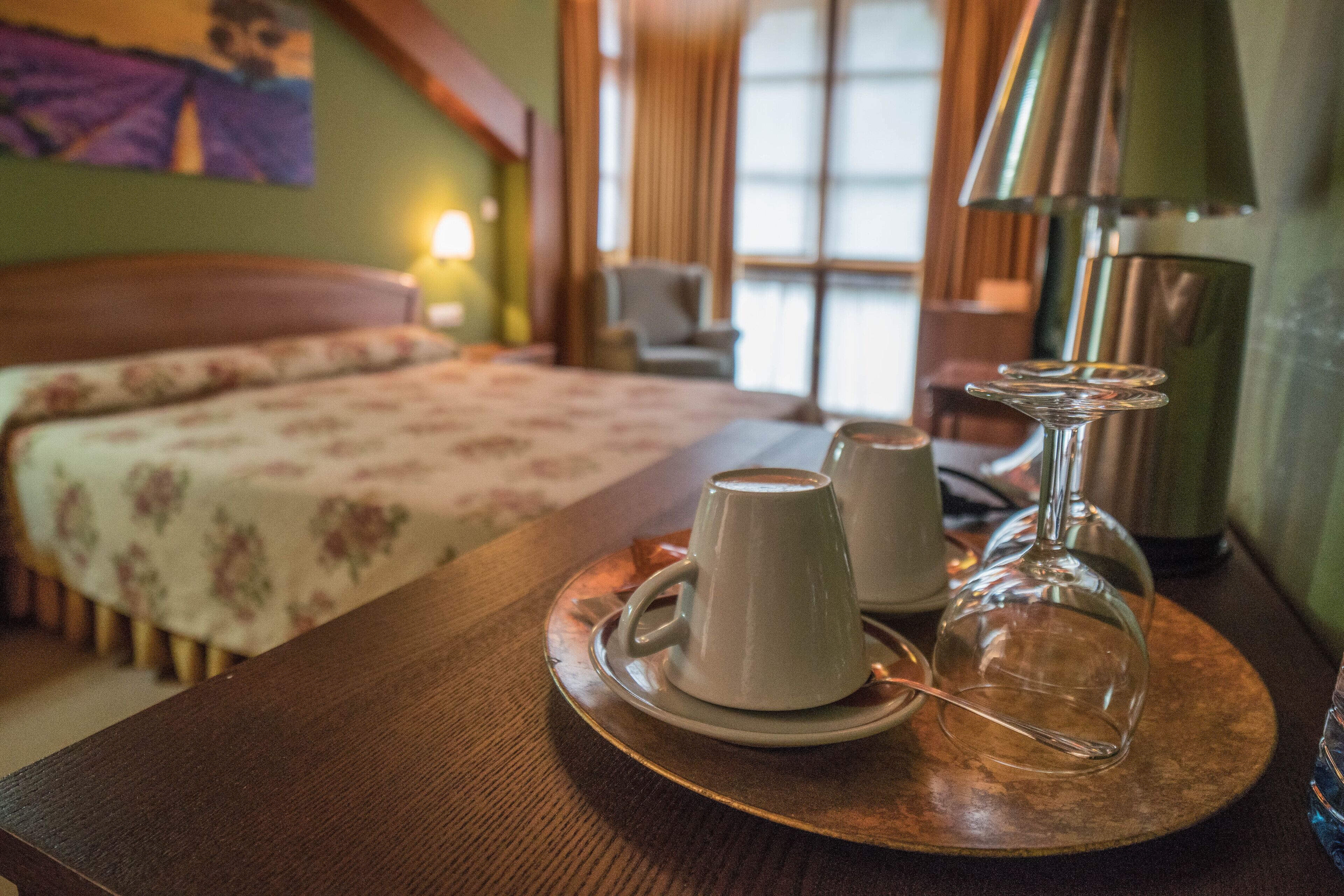 A hotel room with a bed, a table with two cups and a glass, and a lamp.