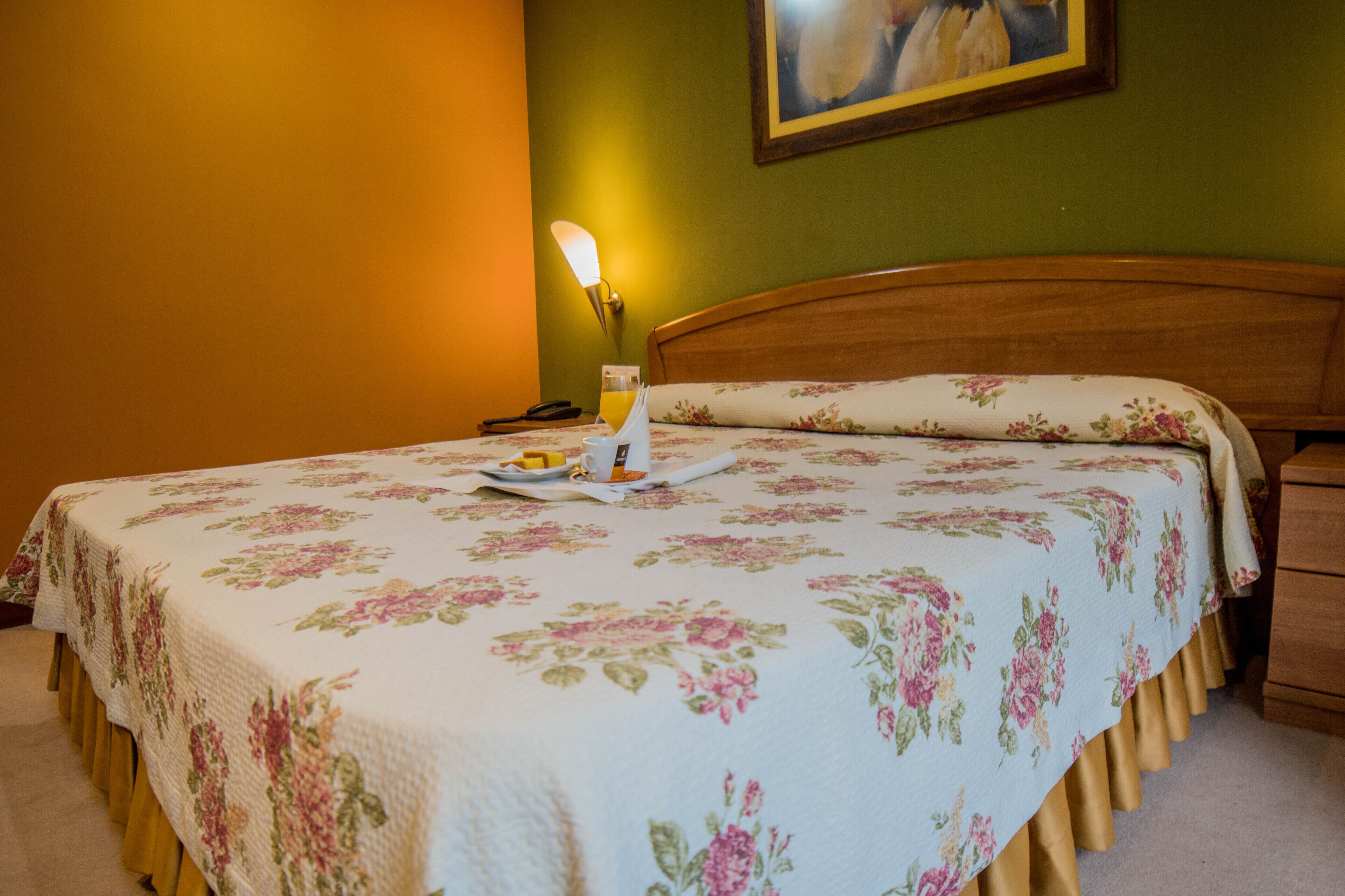 Un lit bien fait, recouvert d’une couvre-lit à motifs floraux, une table de chevet avec une lampe et un tableau accroché au mur.