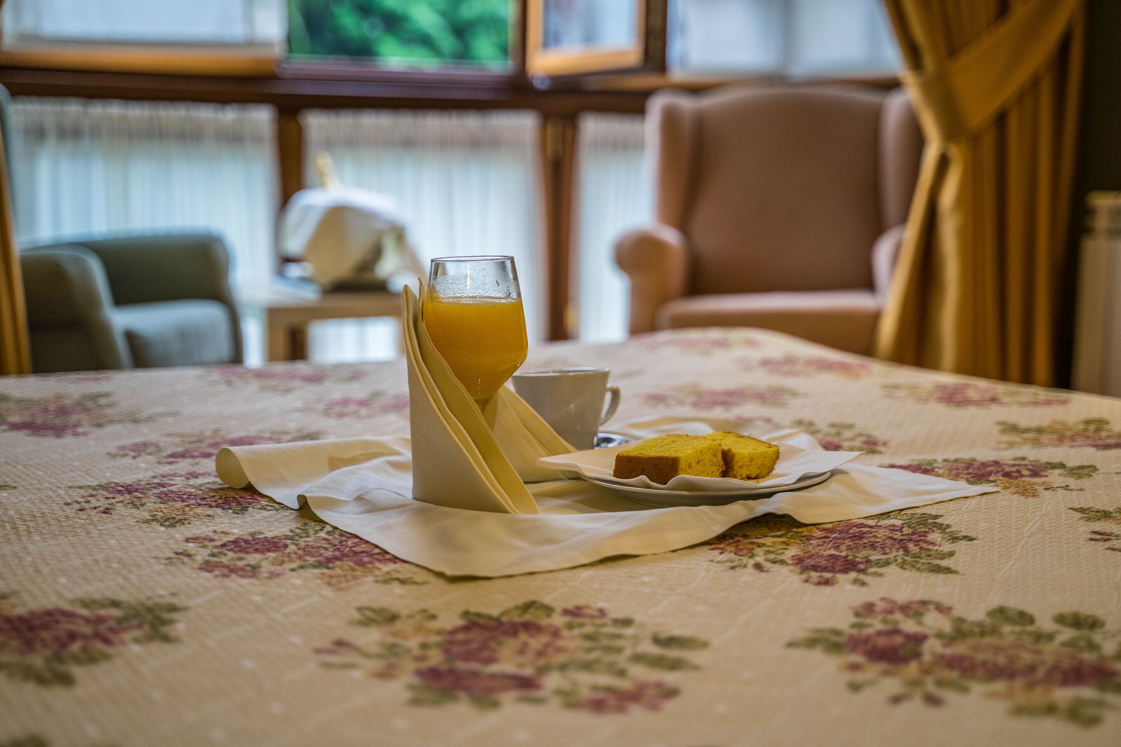 A dining table with a floral tablecloth, a glass of orange juice, a white cup, and a plate with a slice of cake.