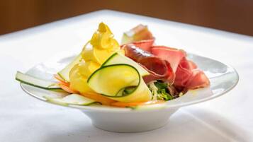 Une assiette blanche contenant une salade composée de concombres et de carottes en tranches, ainsi qu’un morceau de viande rouge, peut-être du prosciutto, le tout posé sur une nappe blanche.