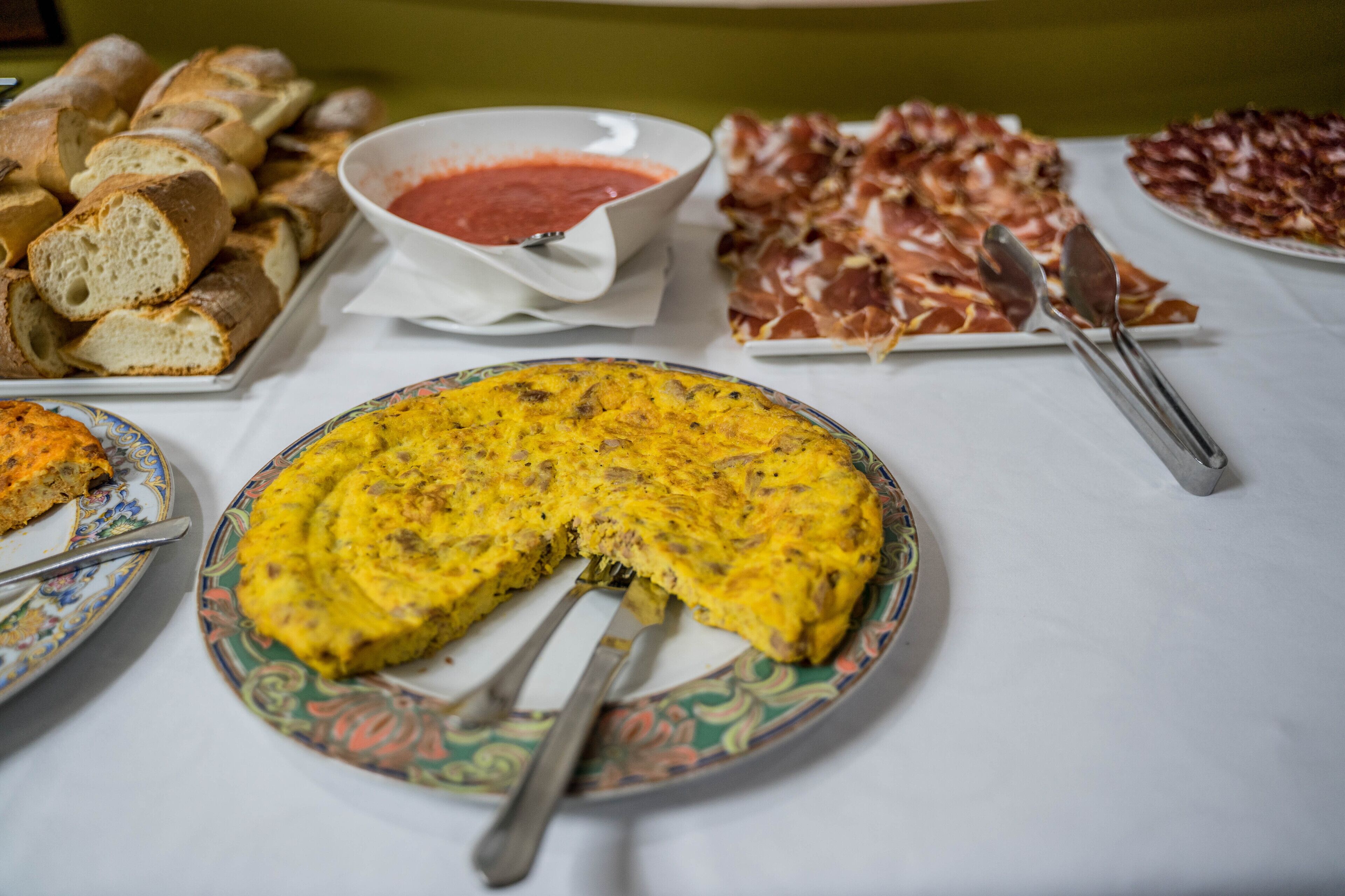 Una mesa con diversos alimentos, como pan, un tazón de salsa roja, un plato de embutidos y una porción de quiche.