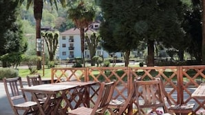 Une terrasse extérieure avec des tables et des chaises en bois, entourée d’une végétation luxuriante et offrant une vue sur un bâtiment en arrière-plan.