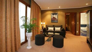 Une chambre d’hôtel avec un canapé, deux poufs, une plante en pot, une lampe et une affiche au mur.