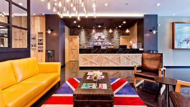 Un hall d’entrée moderne avec un comptoir d’accueil en bois, un canapé jaune et une table ornée d’un chemin de table à motifs Union Jack.