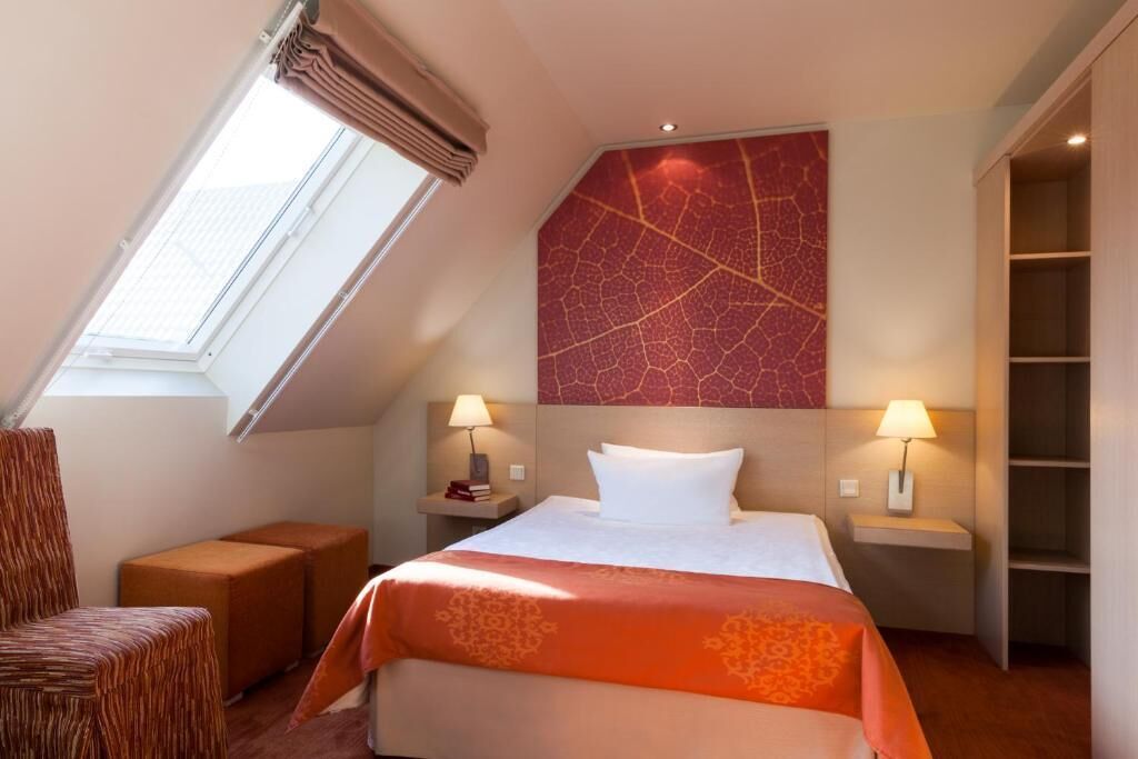 A hotel room with a bed, two bedside lamps, a small table with a lamp, a chair, and a large window.