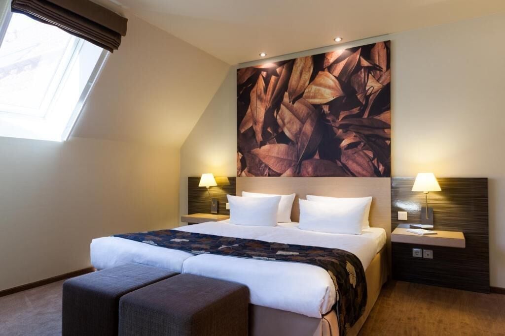 A hotel room with a large bed, two bedside tables with lamps, a headboard with a leaf pattern, and a window with curtains.