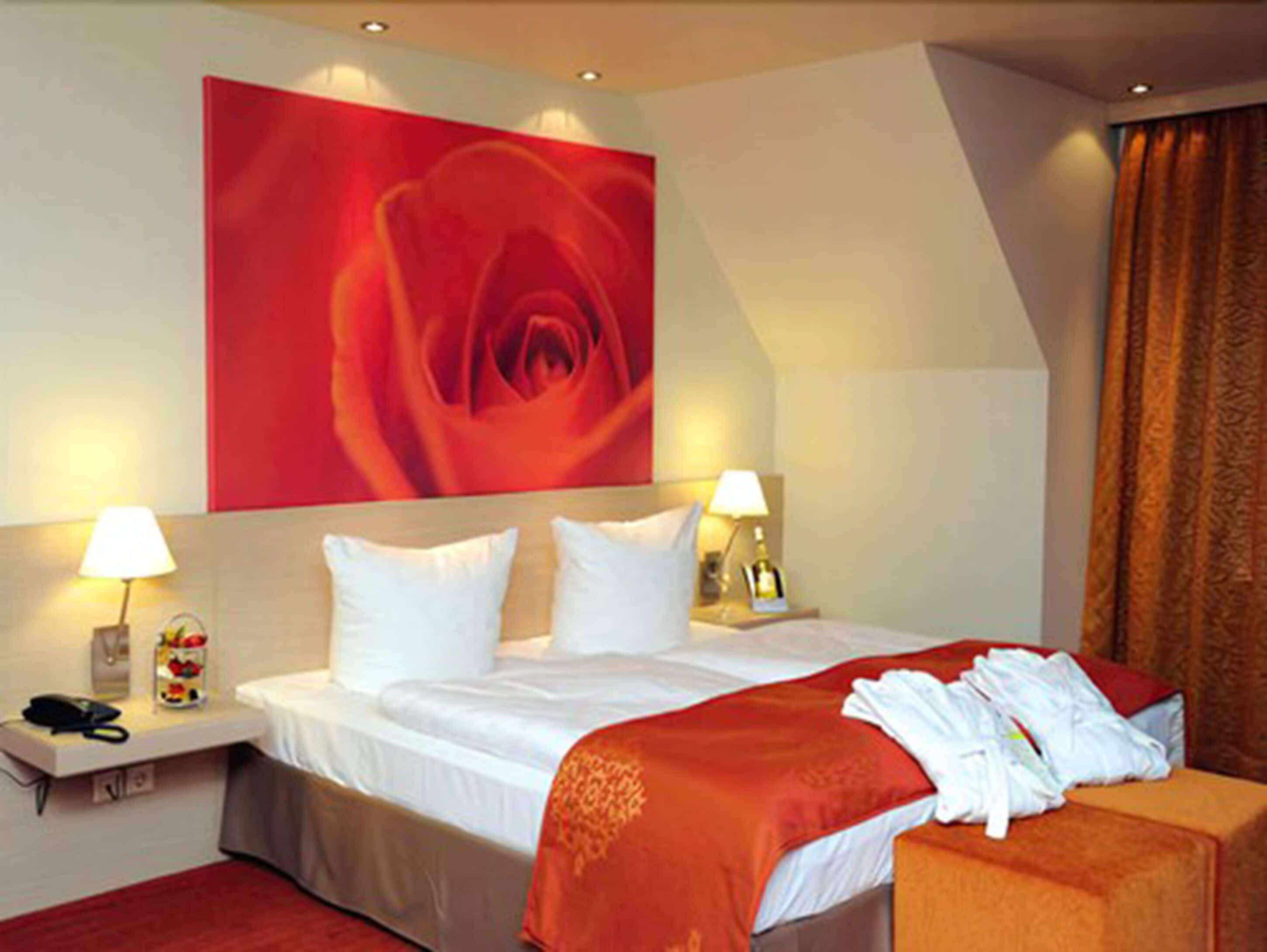 A hotel room with a large red rose painting, a neatly made bed with white linens, two bedside lamps, and a red throw blanket.