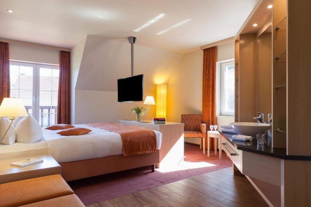 Habitación de hotel con cama, mesita de noche, lámpara, silla, escritorio y baño con lavabo y espejo.